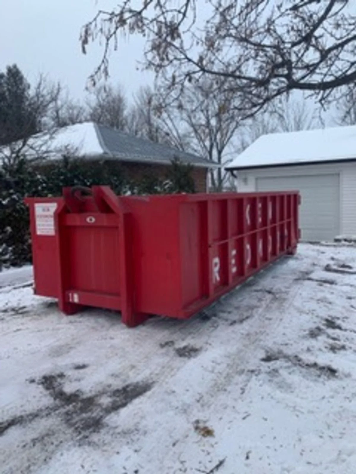 Red dumpster in a snowy driveway with trees and houses in the background.