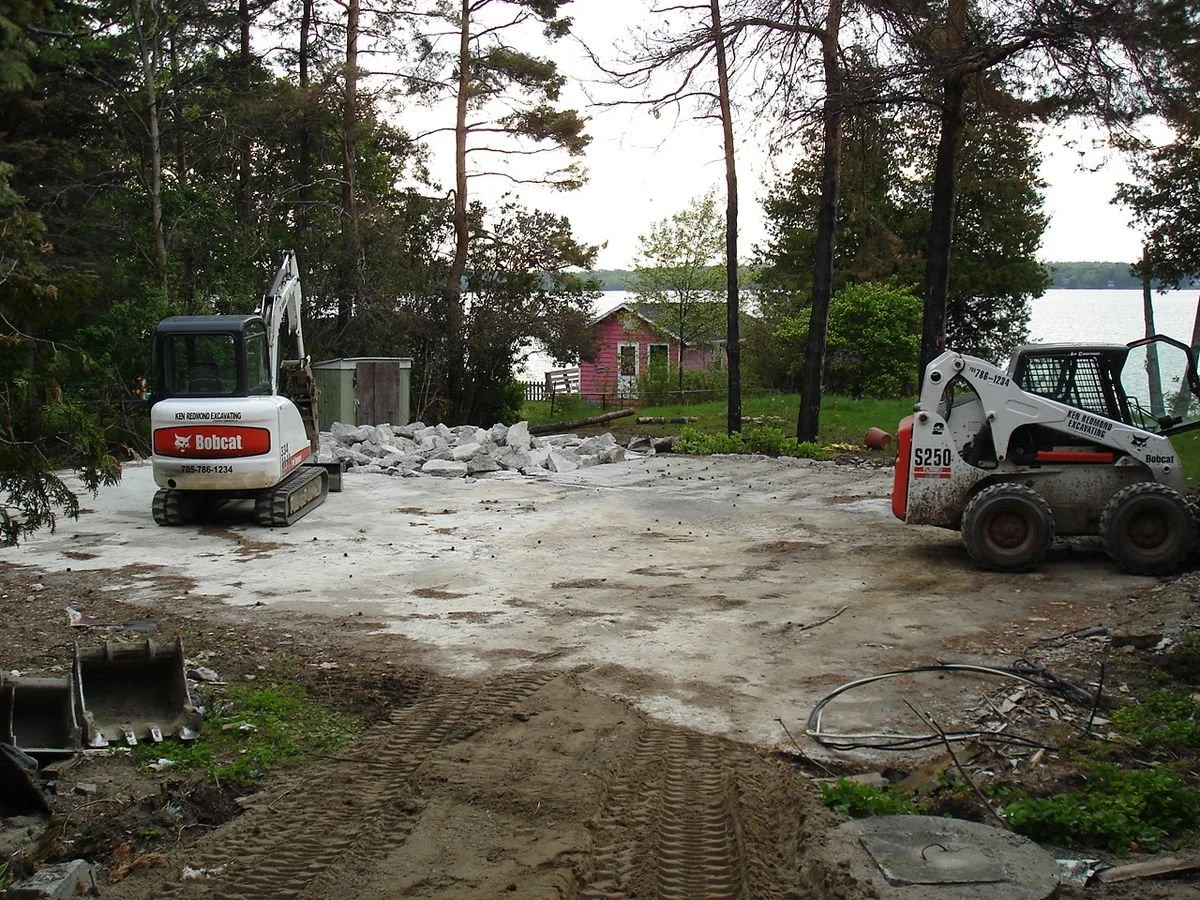 Construction site with two Bobcat excavators, rubble, and disturbed ground near a lake, with a pink house and trees in the background.