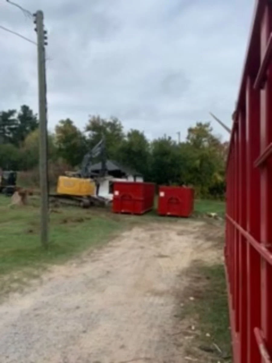 Burned out small building with damaged roof surrounded by trees and red containers, viewed from a gravel path, overcast sky.