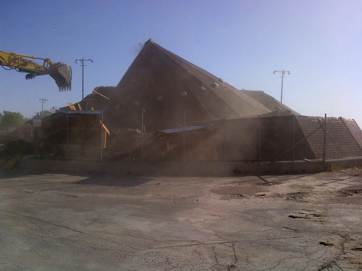 A building collapsed, with debris and dust in the air, a construction excavator arm visible on the left side.