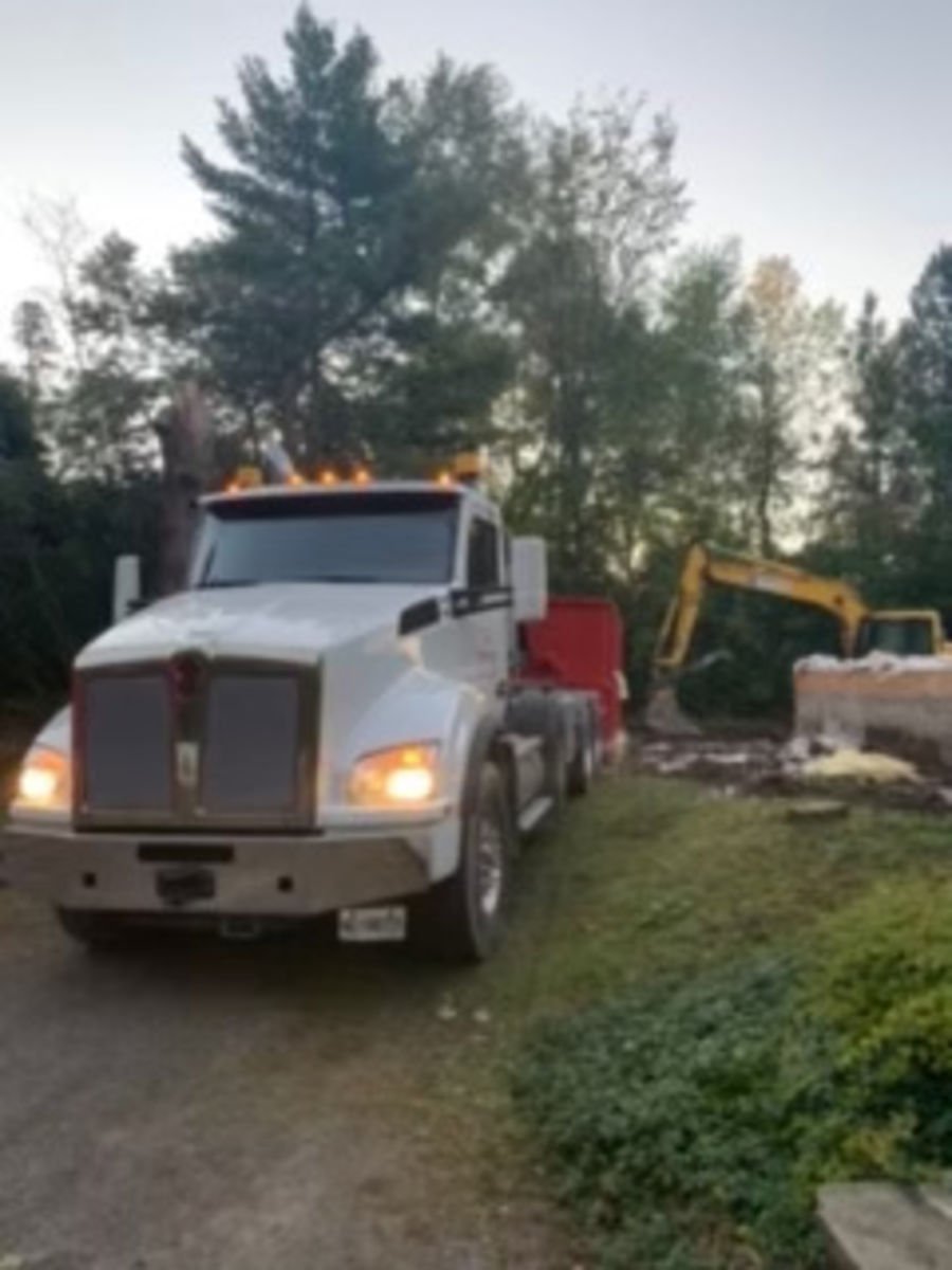 A white semi-truck parked on a grassy area with an excavator in the background, surrounded by trees.