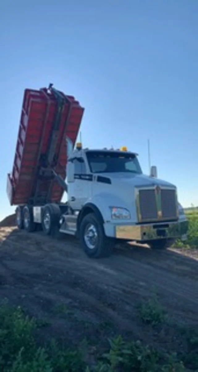 A white dump truck with a red bed tilted upwards on a dirt mound, against a clear blue sky.