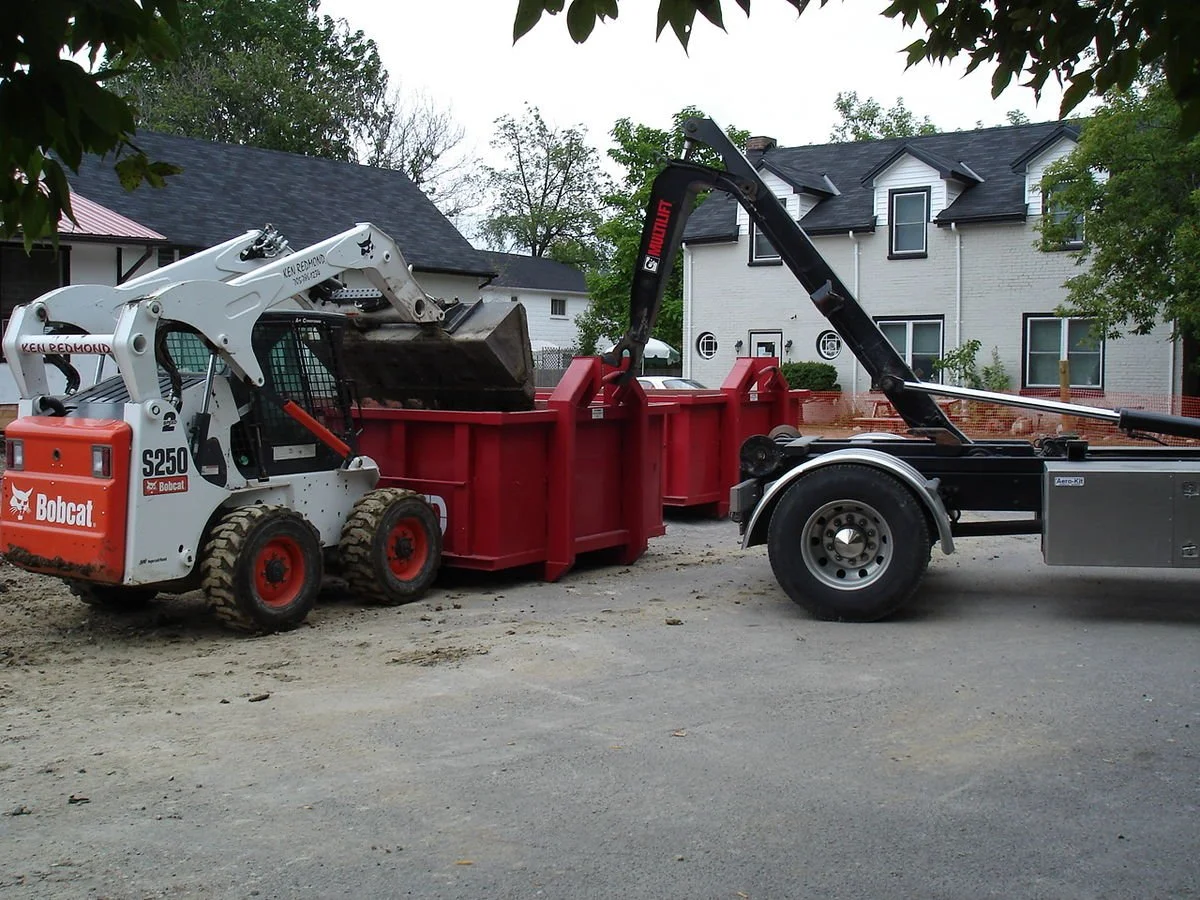 Construction site with a Bobcat compact loader transferring a load of dirt into a red dump trailer connected to a black tow truck.
