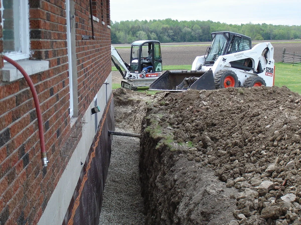 Construction site with a trench dug alongside a brick house and two skid steer loaders working in the background.