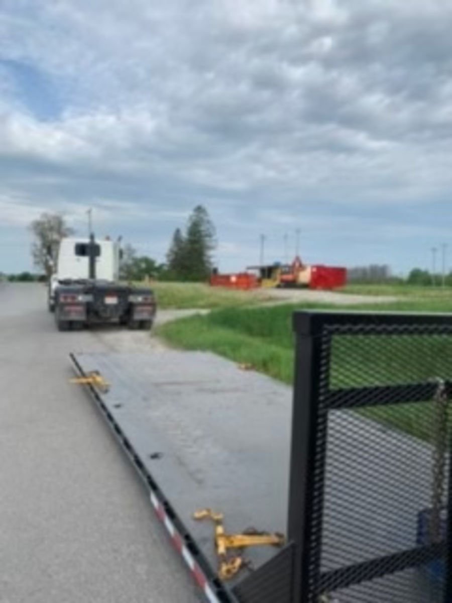 A flatbed trailer attached to a pickup truck parked on a rural road, with construction equipment and trailers in the background under a cloudy sky.
