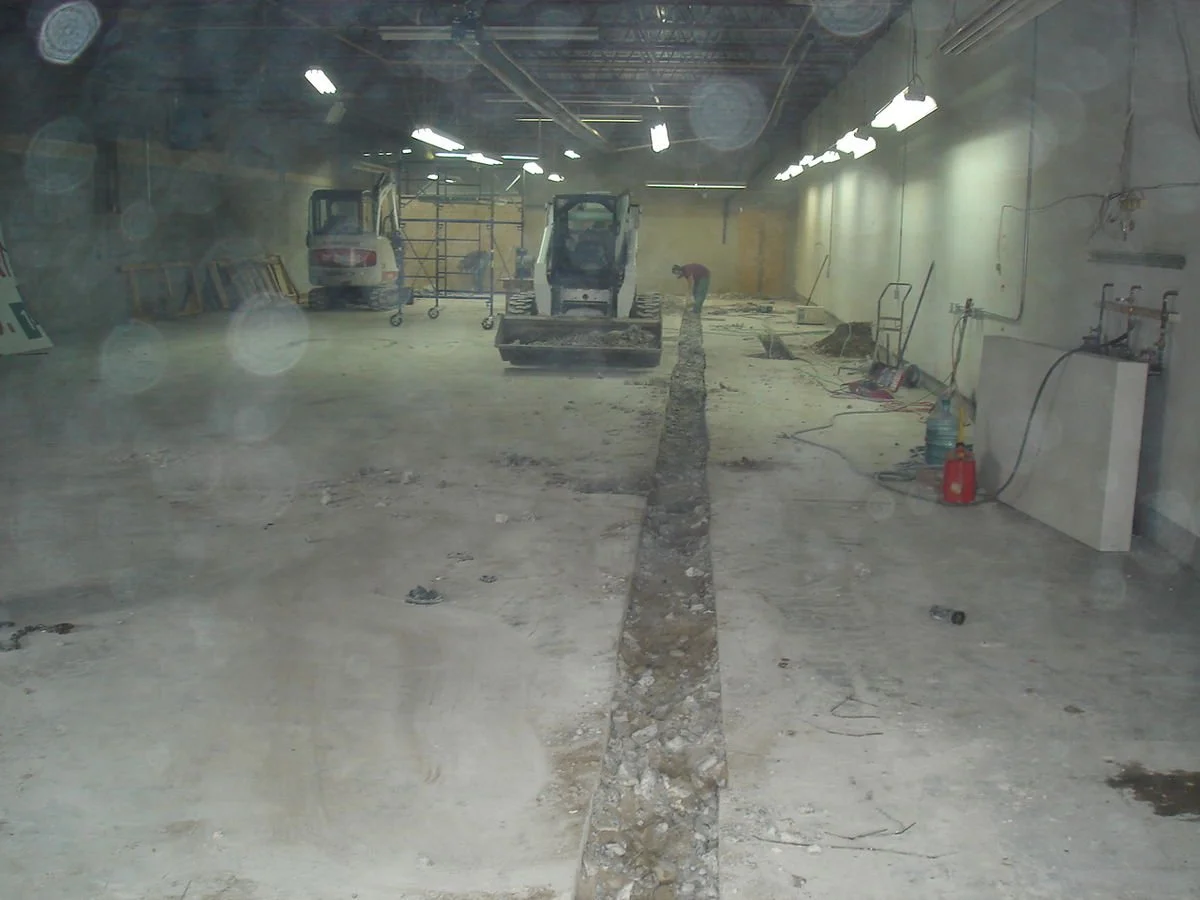Construction site inside a building with machinery and workers working on floor trench.