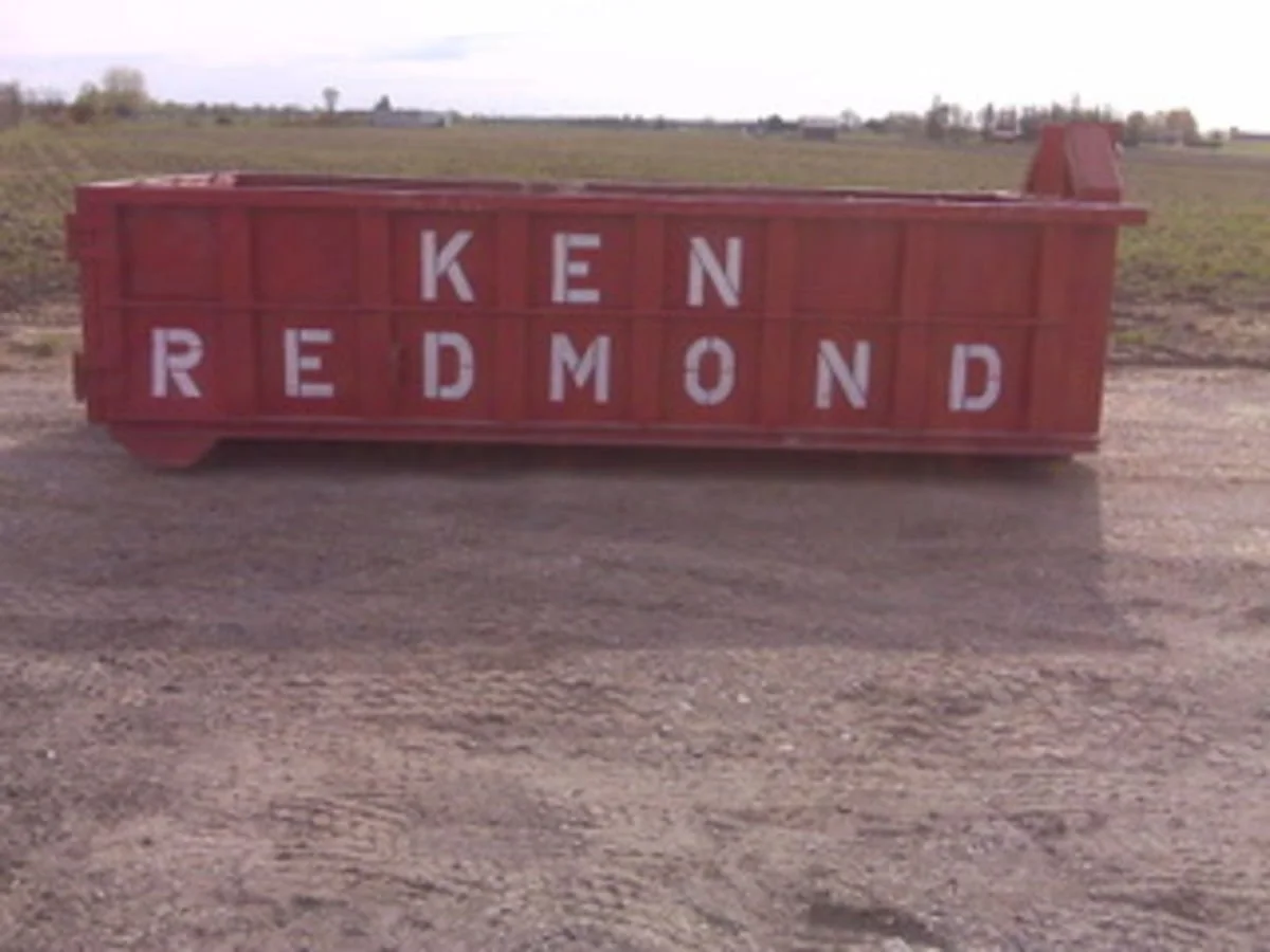 Red barn with white letters spelling 'KEN REDMOND' on the side, situated on a dirt road in a rural area.