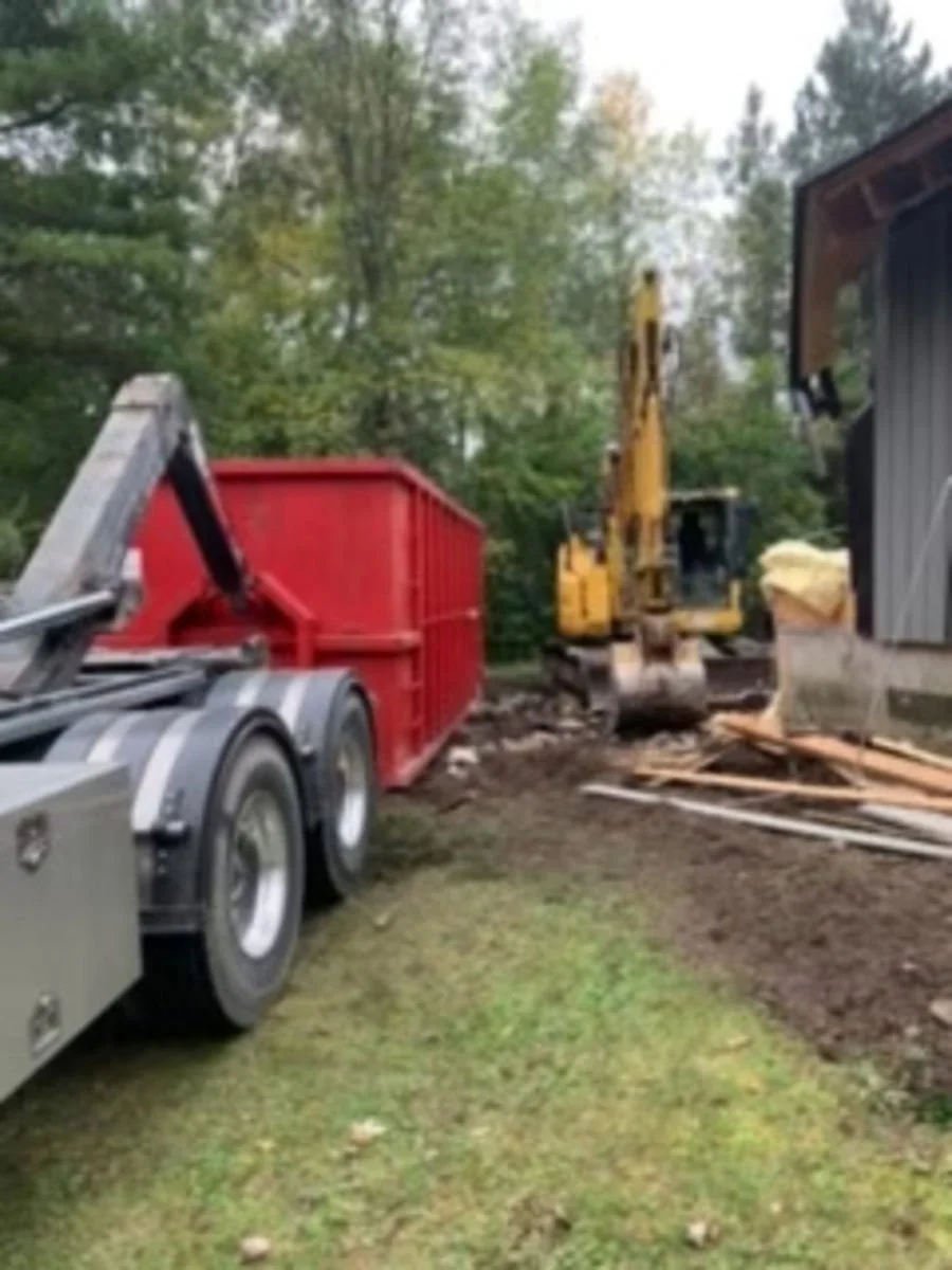 Construction site showing a yellow excavator, a red dumpster, and wooden planks near a building with trees in the background.