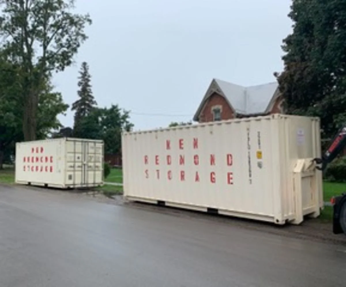 Two large white storage containers on the side of a street, marked with red letters reading "KEW STORAGE" and "INDUSTRIAL STORAGE," with trees and a brick house in the background.