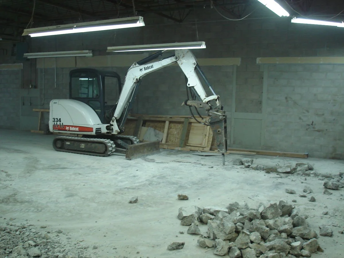 A white Bobcat mini excavator clearing a concrete floor with rocks and debris in an indoor construction site.