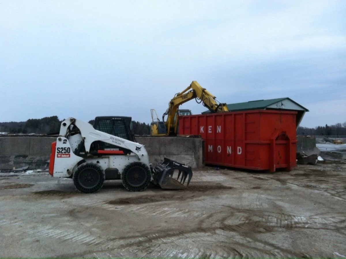Construction site with a Bobcat skid-steer loader in the foreground and a yellow excavator loading dirt into a red dumpster with the words 'KEN MUND' on it, set against a cloudy sky.