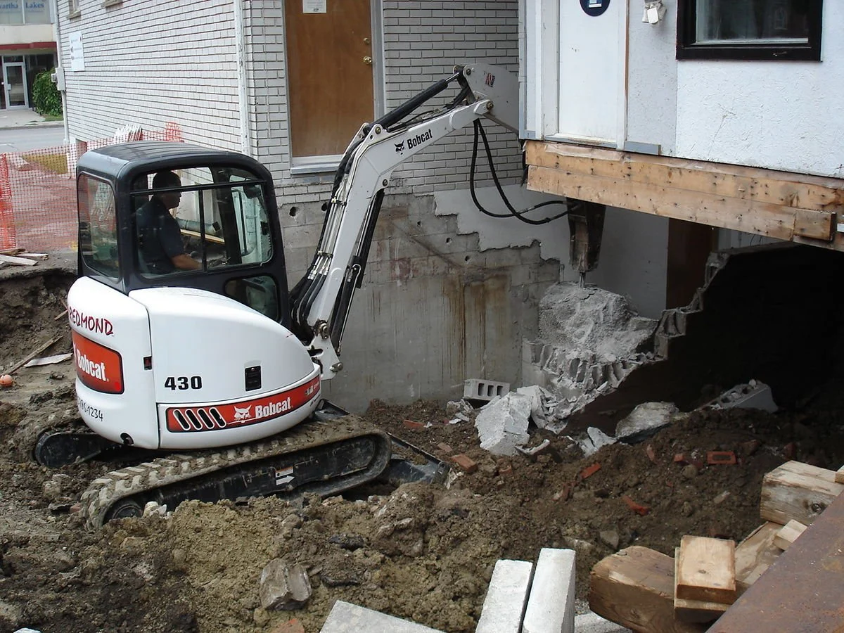 A small white and black Bobcat excavator working on construction, demolishing part of a building foundation, with dirt, bricks, and debris around.