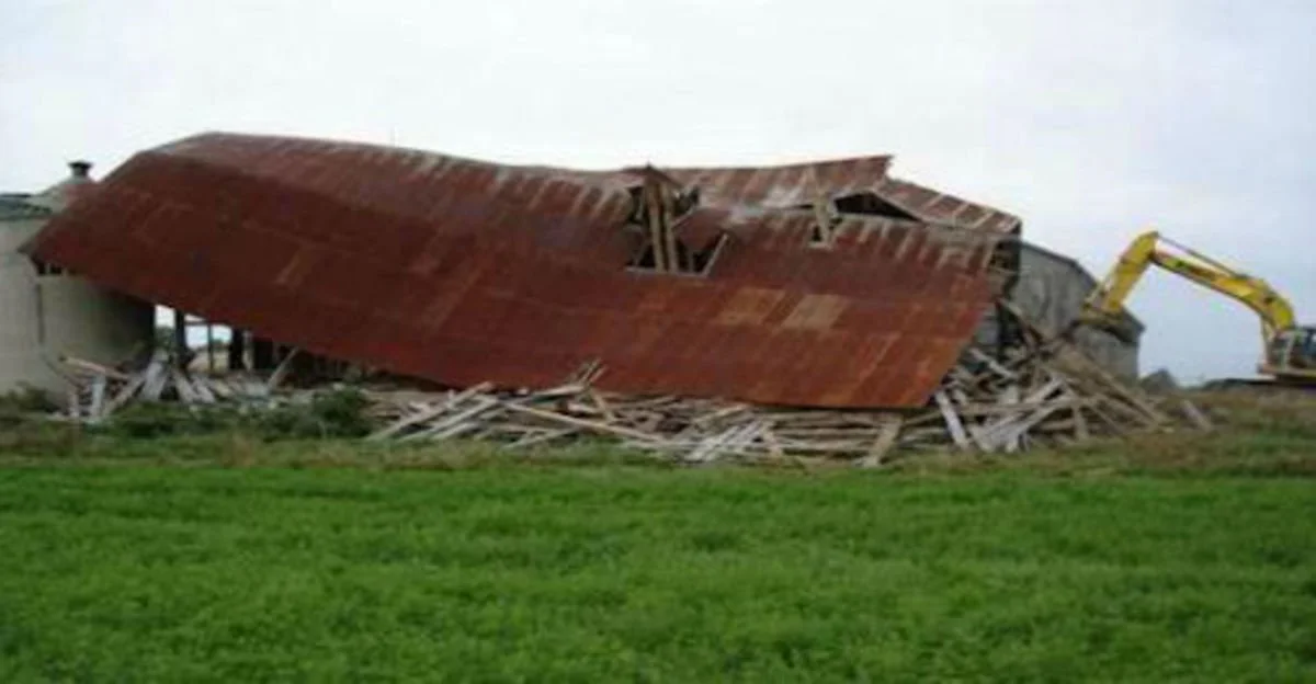 A partially collapsed building with a large, rusted metal roof, supported by wooden beams, with construction equipment nearby.