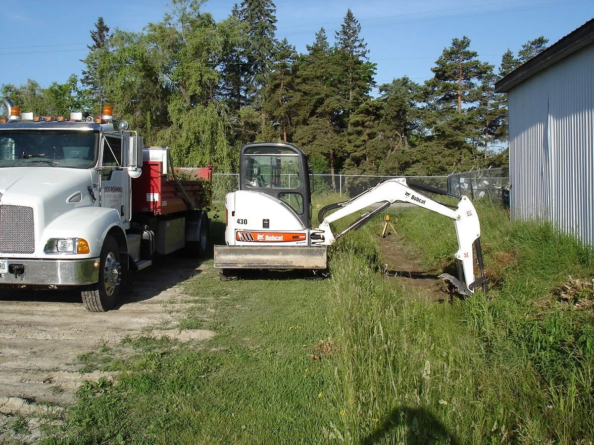 A white Bobcat mini excavator is parked next to a large white utility truck on a grassy area with trees in the background.