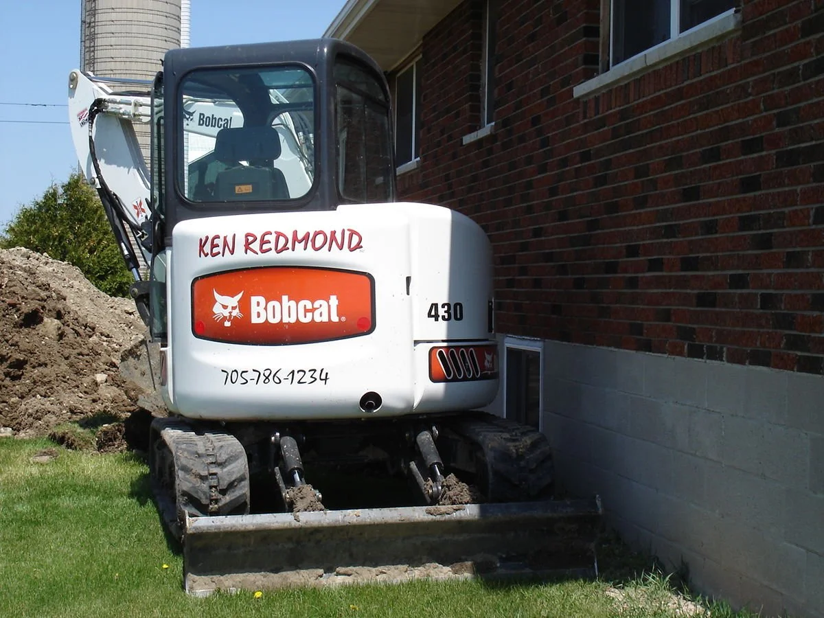 A small white Bobcat excavator with a black cab is positioned next to a brick building, with dirt piled nearby and green grass in the foreground.