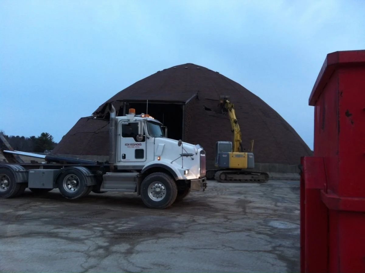 Construction site with a large dome-shaped structure being worked on, a white truck, and a yellow excavator digging into the dome. A red container is visible on the right side.