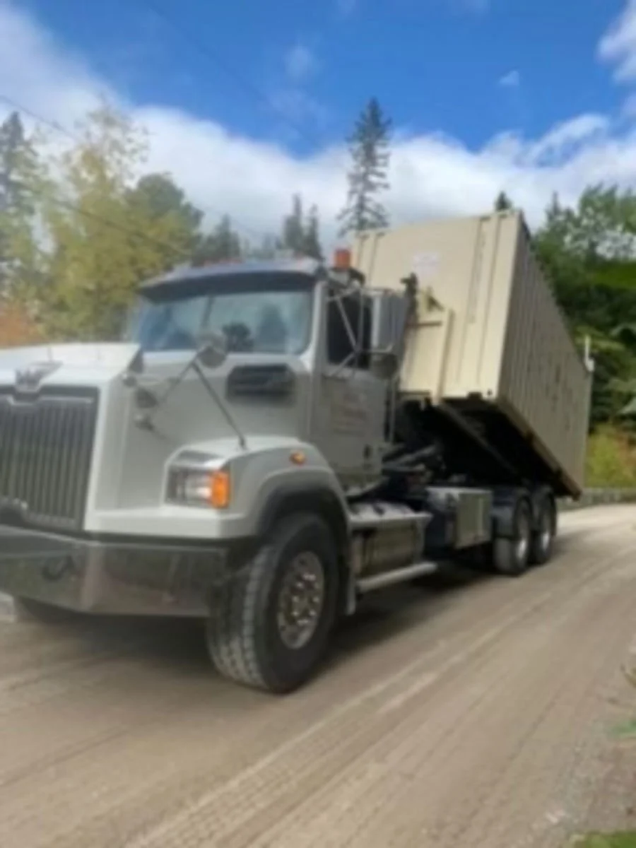 A large gray dump truck with its bed raised, parked on a dirt road surrounded by trees and blue sky.