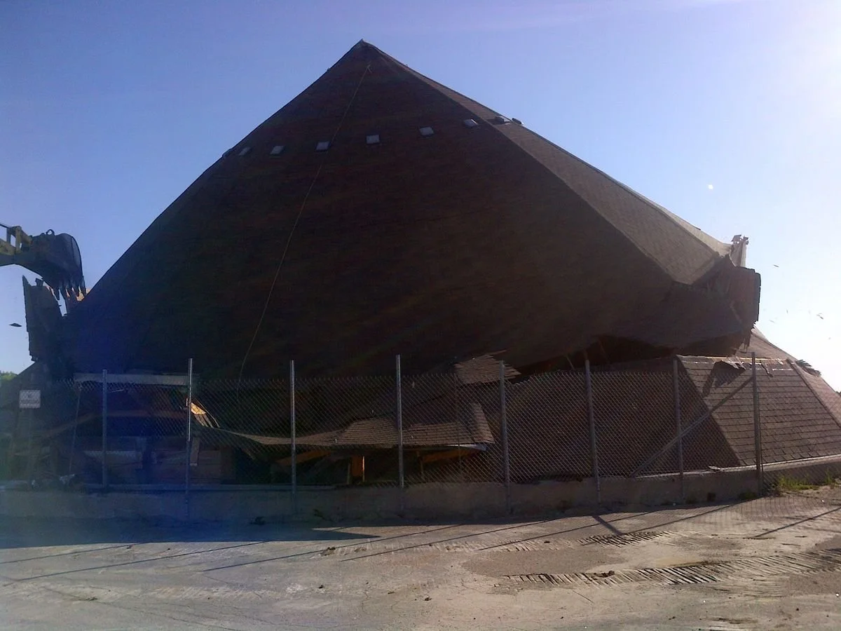 A building featuring a steep, triangular roof that appears to be collapsing or tipping over, surrounded by a chain-link fence and construction equipment.