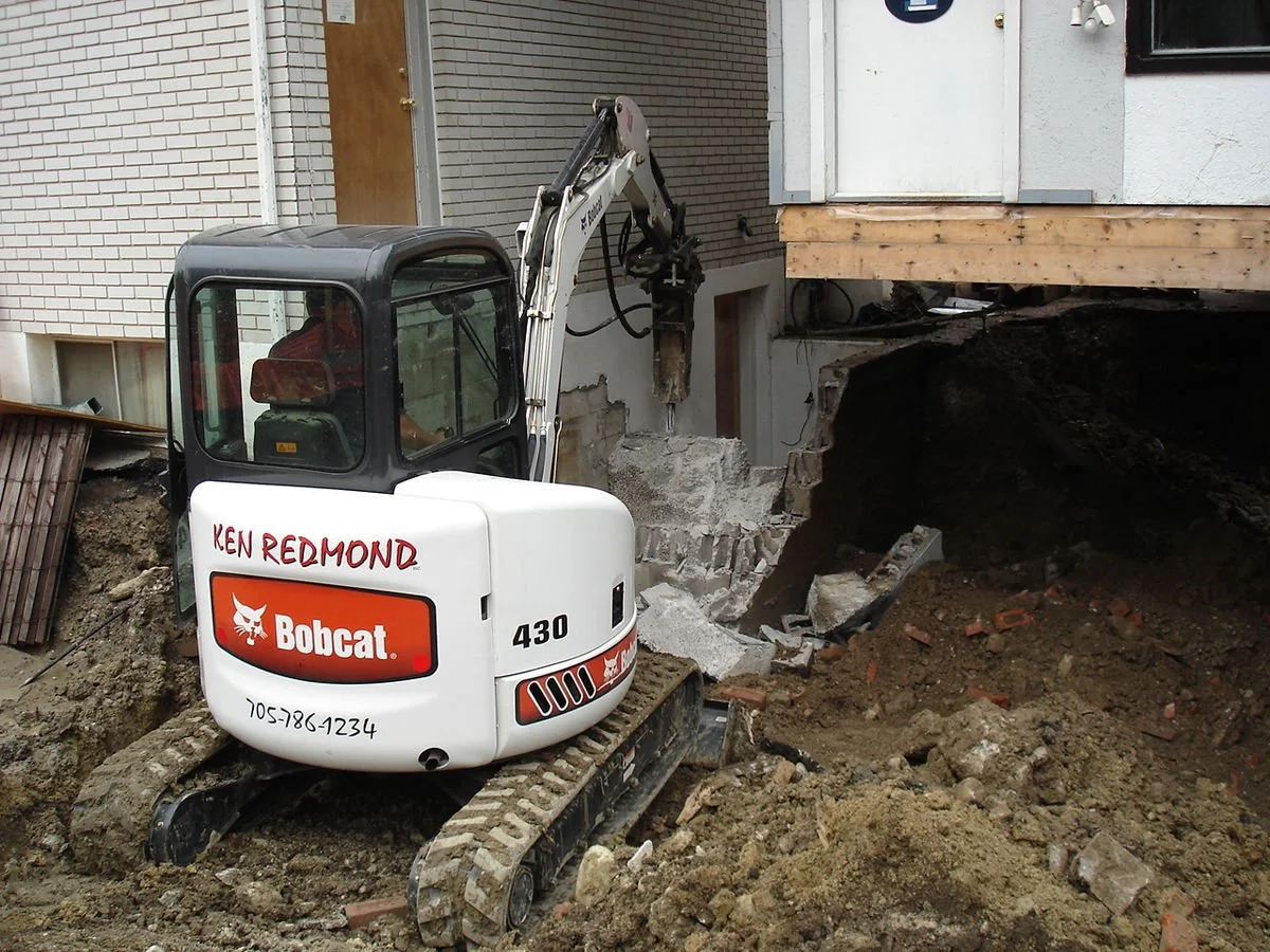 A small bobcat excavator digging near a house foundation with a plywood platform. The house has a white exterior with a brick wall and a door.