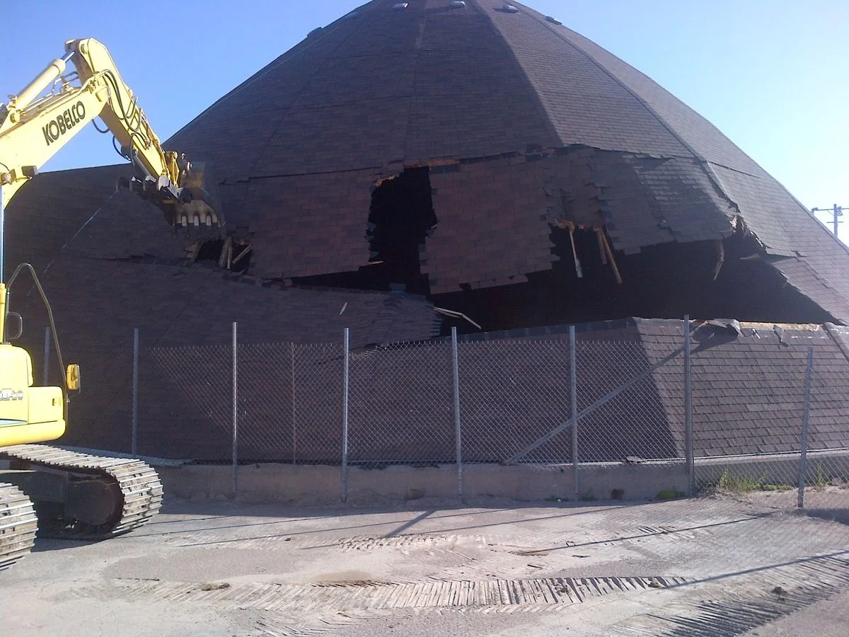 A construction site with a large, damaged, conical building or structure, possibly a dome, with a huge hole in its surface. A yellow excavator is working on the damages, and a chain-link fence surrounds the area.