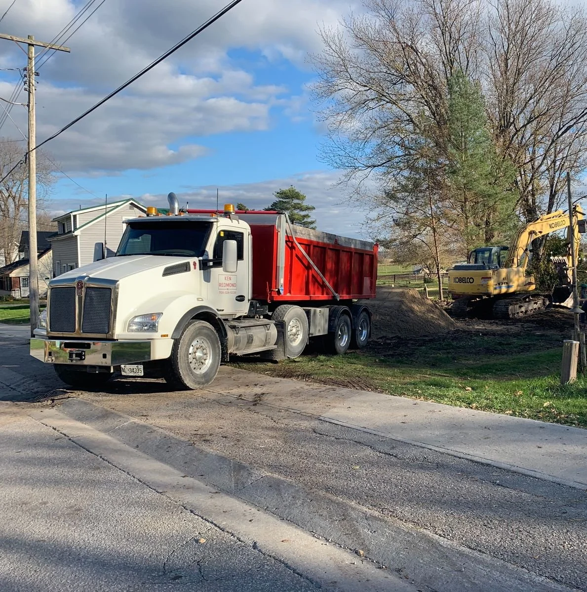 A white dump truck with a red bed parked on a residential street, with a yellow excavator nearby digging into the ground among trees and houses, under a partly cloudy sky.