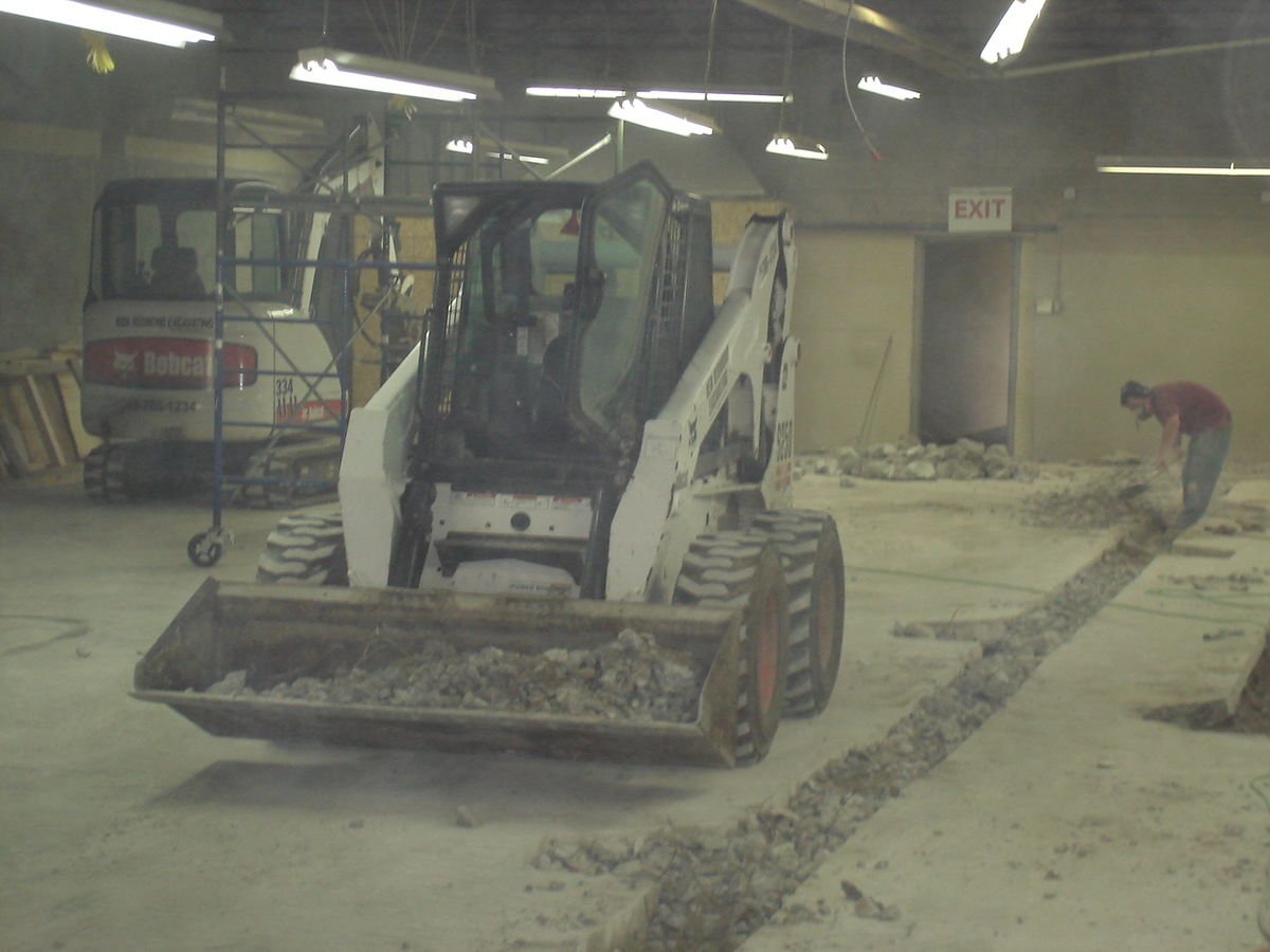 Indoor construction site with a skid-steer loader scooping dirt, a worker digging, and construction equipment.