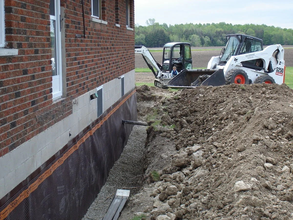 Construction workers use a skid steer loader to dig a trench next to a brick house foundation, with new gravel and drainage materials being installed.