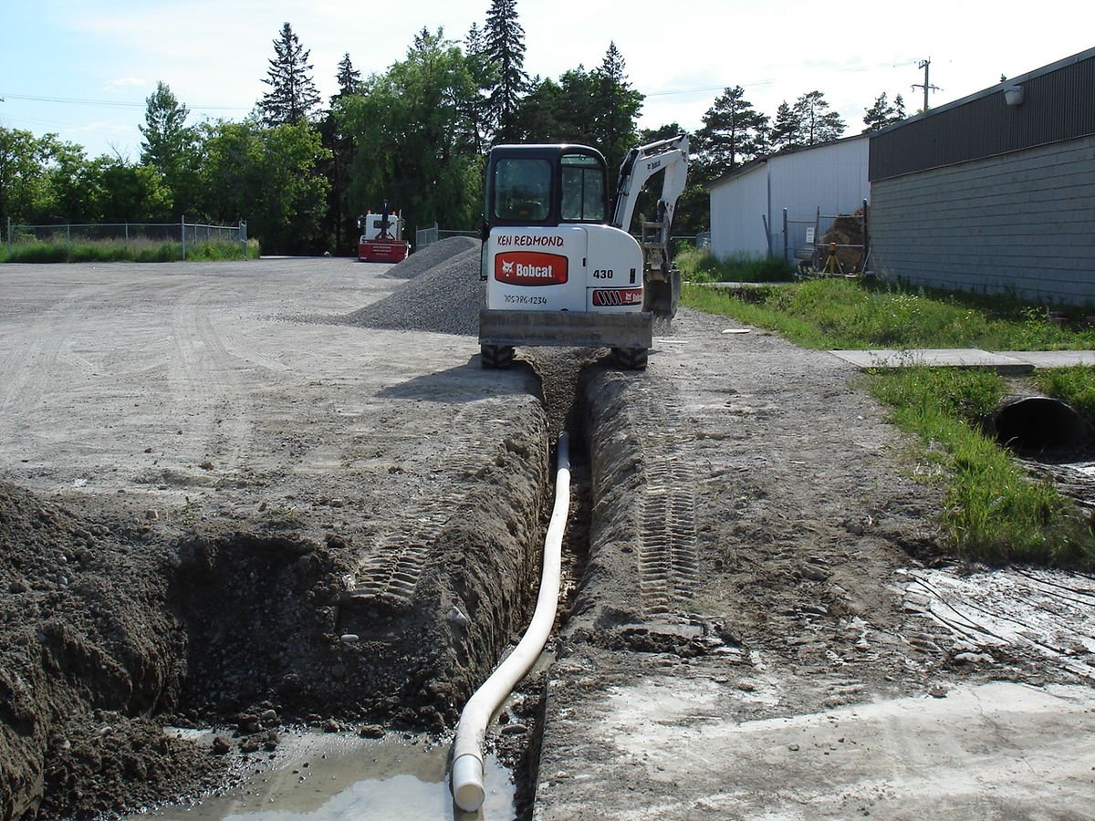 Construction site with a mini excavator working in a trench, pulling a white pipe, with a pile of gravel and a red machine in the background.