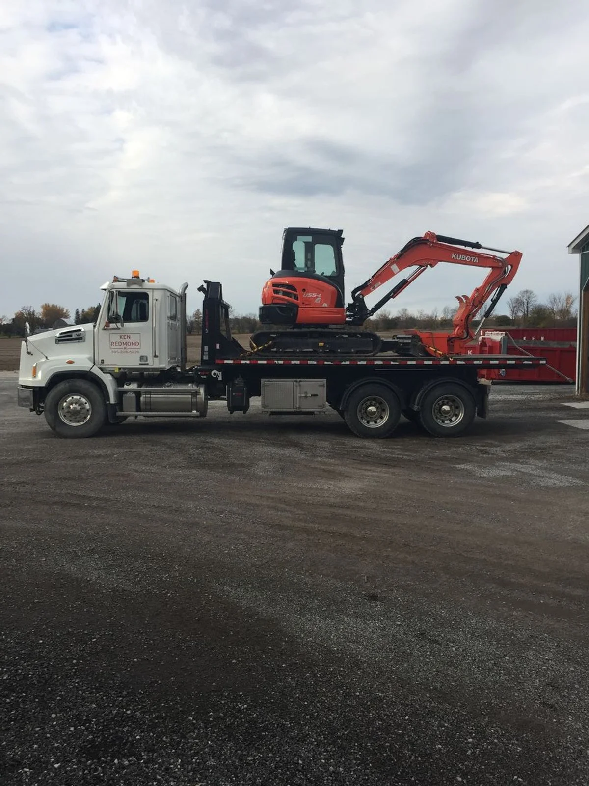 A white flatbed truck carrying an orange mini excavator on a gravel surface with a cloudy sky above.