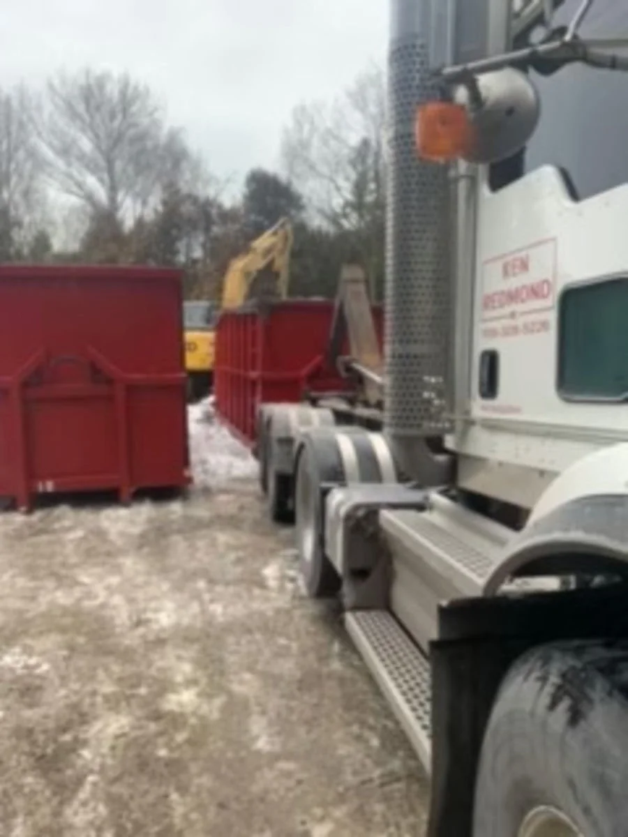 A white semi-truck parked next to red dumpsters on a snowy ground, with a construction excavator in the background during overcast weather.