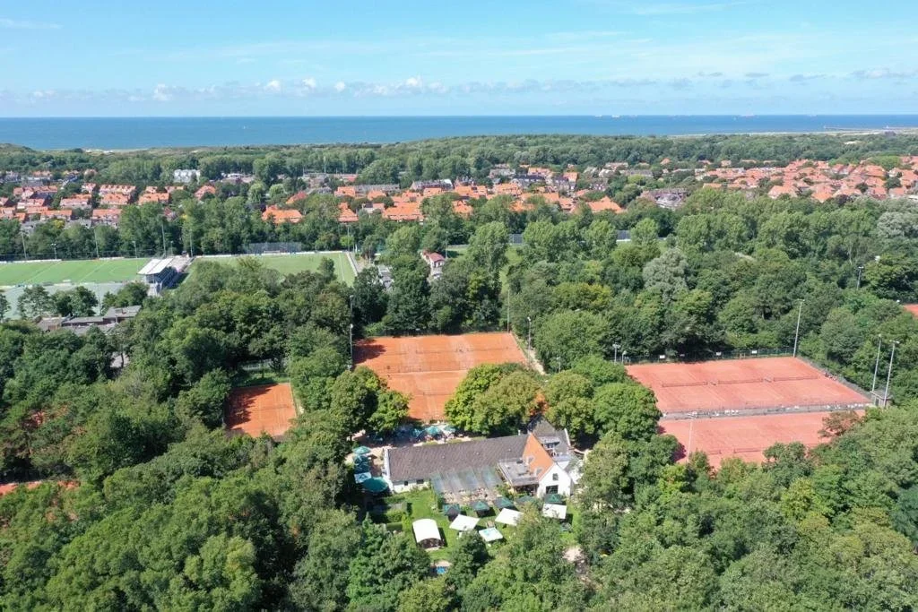 Aerial view of a green area with tennis courts, a house, tents, trees, and a residential neighborhood with orange rooftops near the coastline.