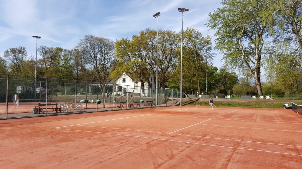 A tennis court with red clay surface, surrounded by a chain-link fence, with a person walking on the court. There are large trees and a white house in the background, with tall light poles and a blue sky overhead.
