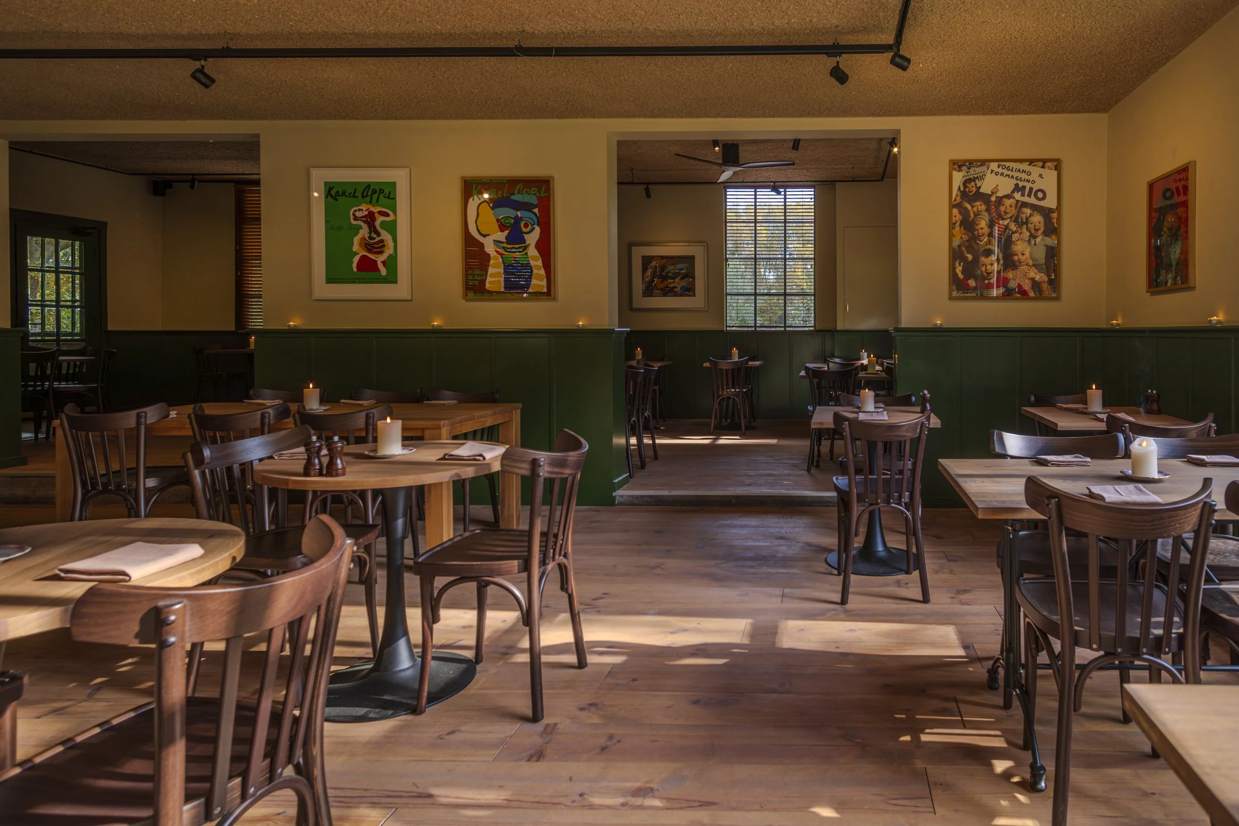 Empty restaurant interior with wooden tables and chairs, lit candles, green paneled walls, colorful art posters, and sunlight streaming through windows.
