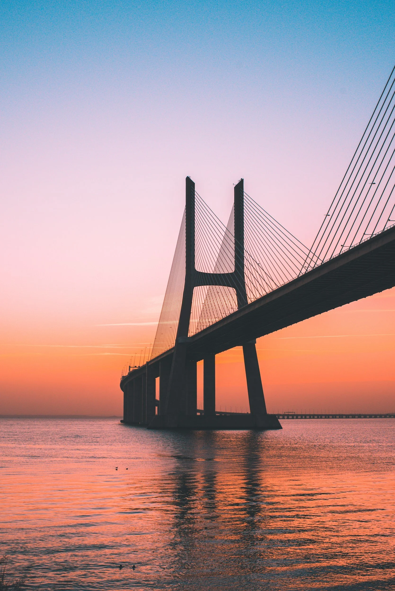 A suspension bridge over water during sunset with a colorful sky.
