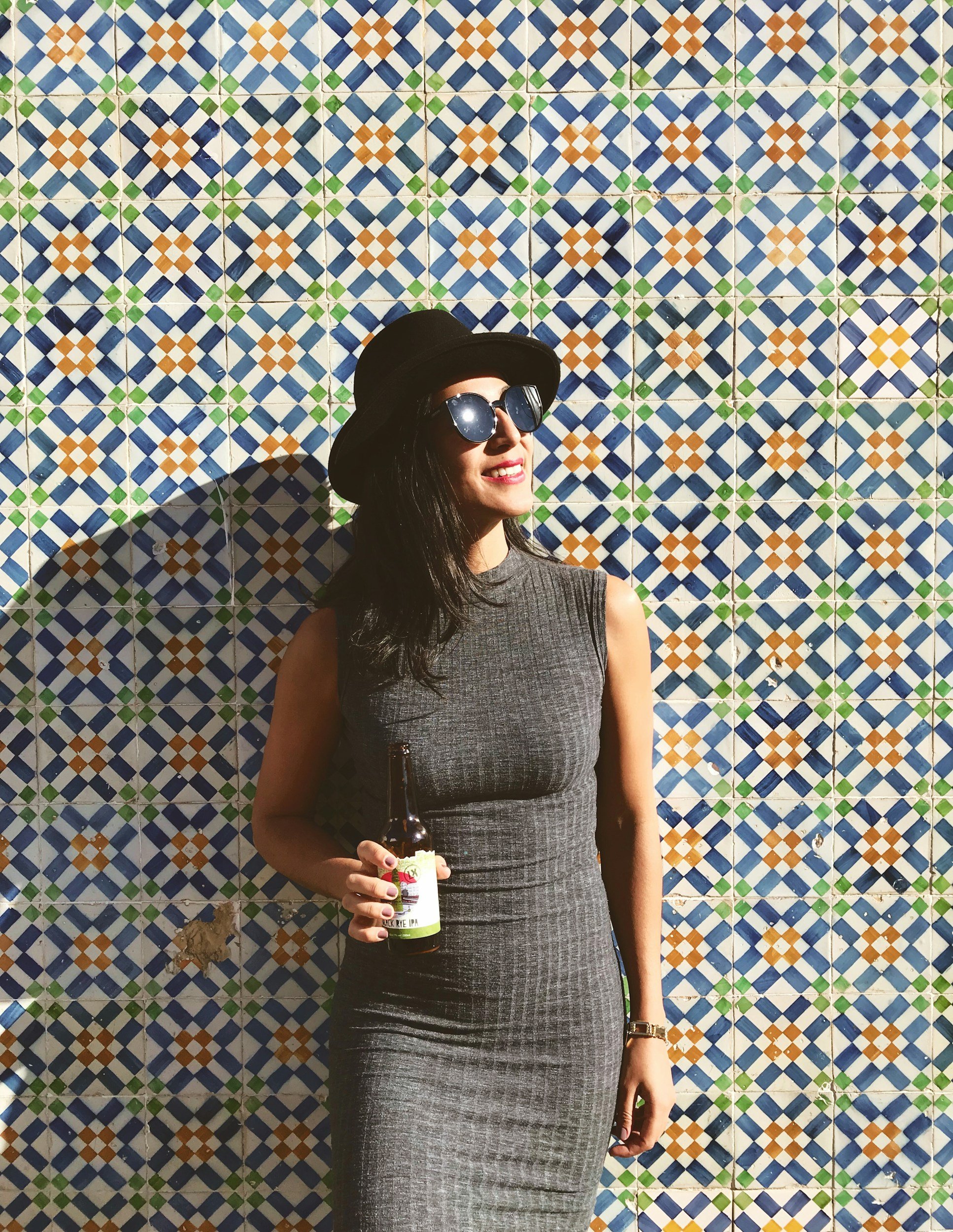 Woman wearing a black hat and sunglasses, holding a beer bottle, standing in front of a colorful tiled wall.