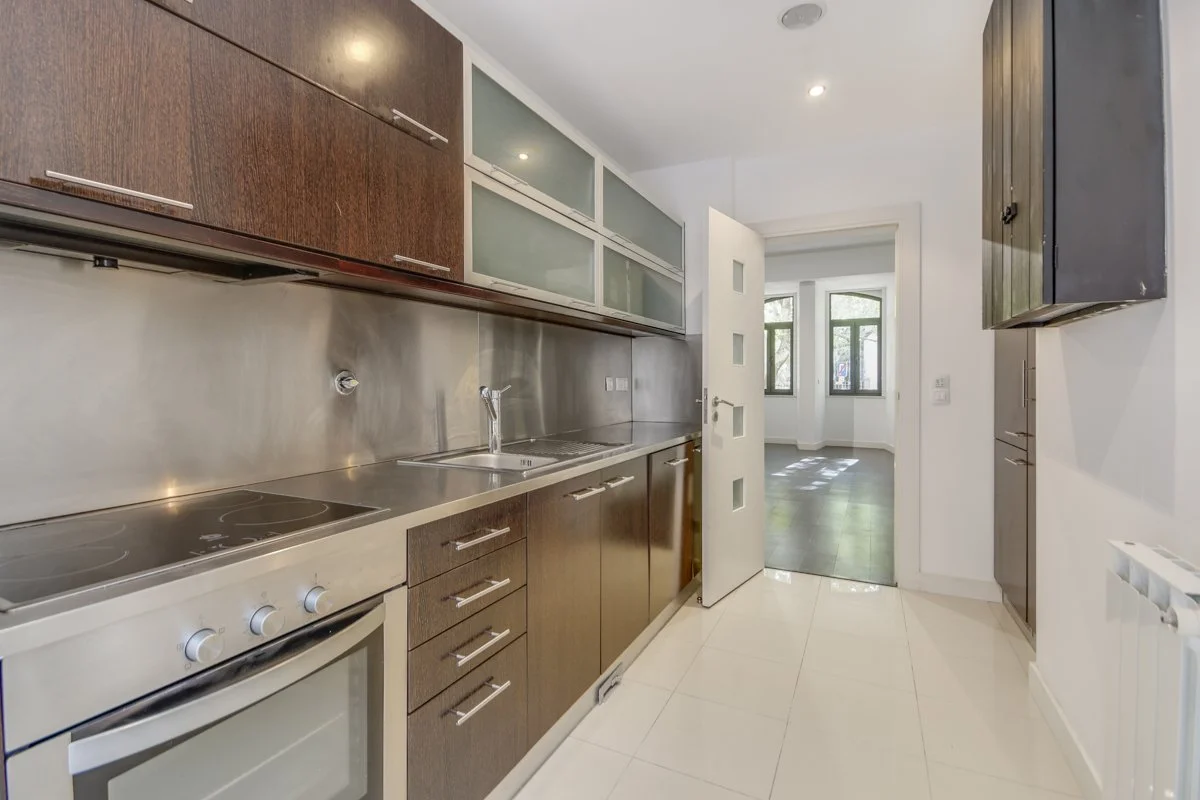 Modern kitchen with dark wood cabinets, stainless steel backsplash, built-in oven, electric stovetop, and a small sink, leading into a bright living area with large windows.