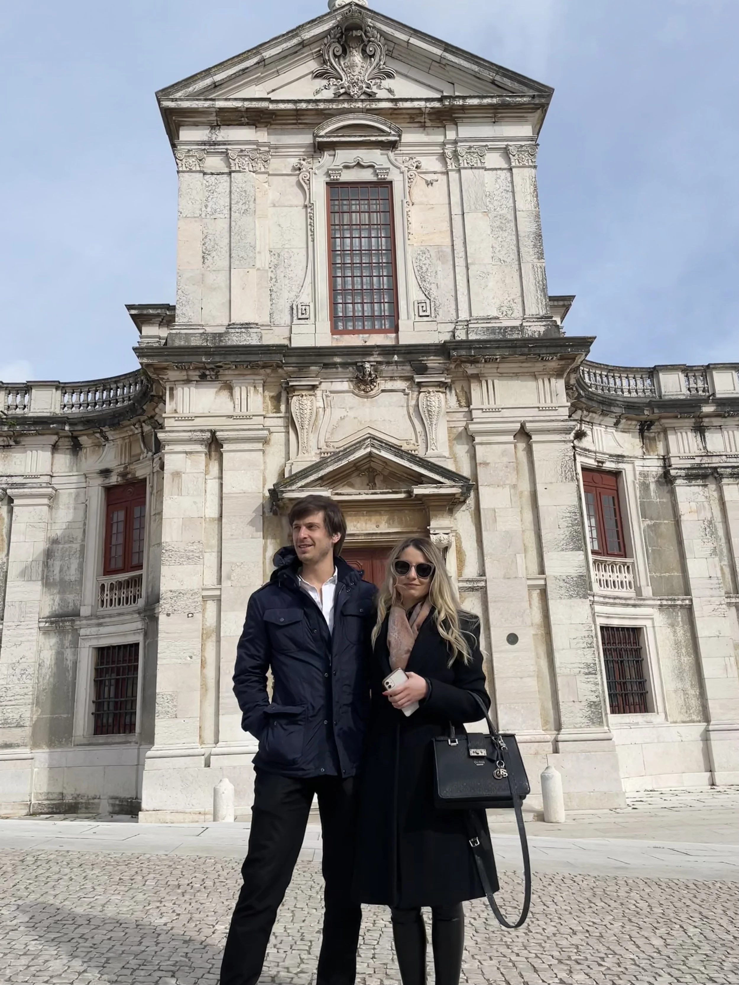 A man and a woman standing in front of a historic stone building with ornate architectural details and red window frames. The man is wearing a dark jacket, and the woman is wearing sunglasses, a black coat, and holding a handbag and phone.