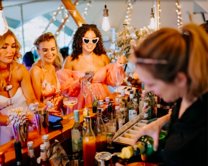 Women enjoying a social gathering at a bar or drinks station with bottles and mixers, one woman wearing heart-shaped sunglasses, others smiling, ambiance with hanging lights and outdoor setting.