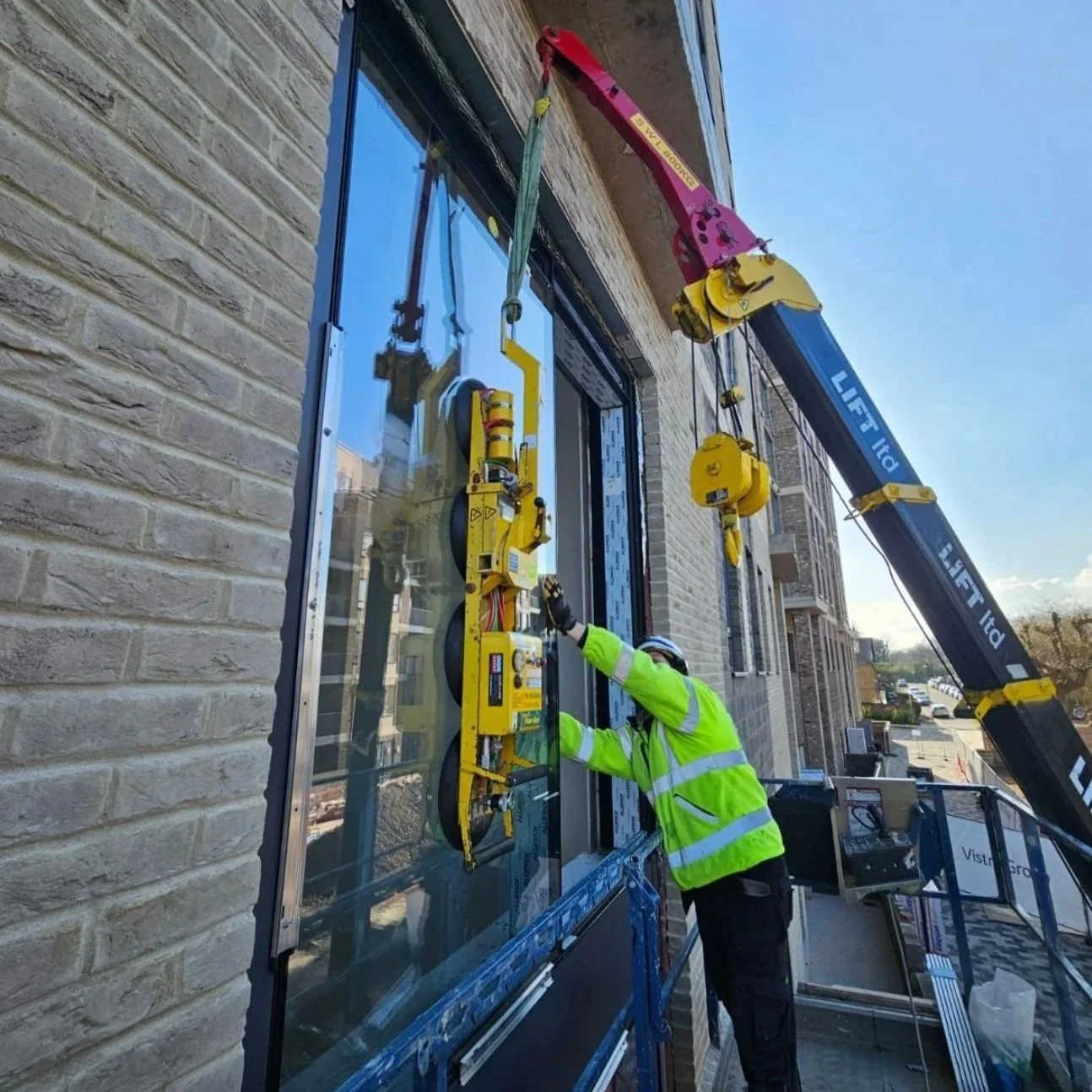 A construction worker in a neon yellow safety jacket and helmet installing or repairing a large glass window on a brick building. A crane stabilizes the glass panel, and the worker is using tools to secure it.