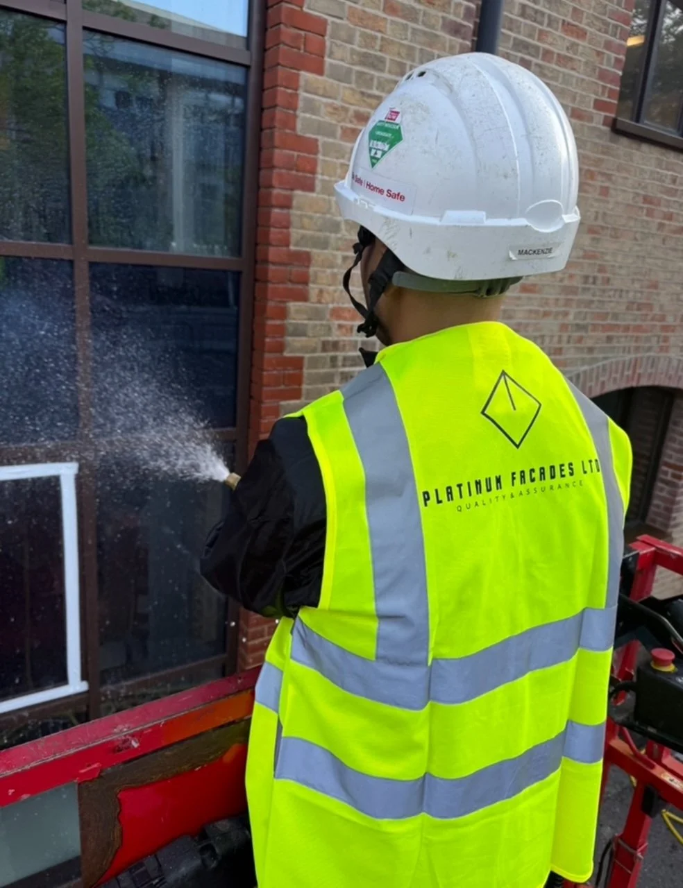 A worker wearing a yellow safety vest and a white helmet using a spray tool on a glass window of a brick building.