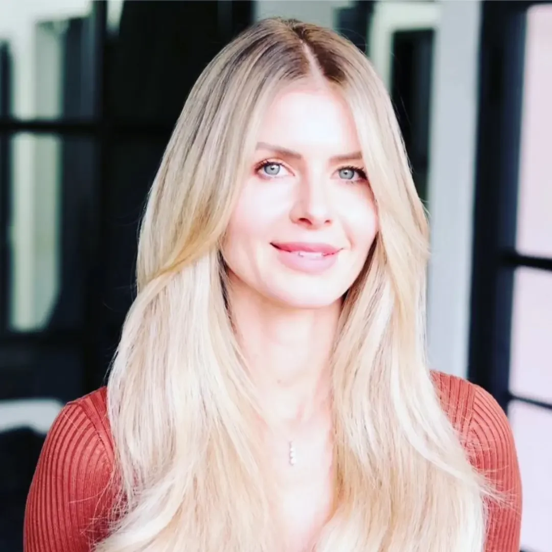 A woman with long blonde hair and blue eyes, wearing a red top, smiling in a modern indoor setting.