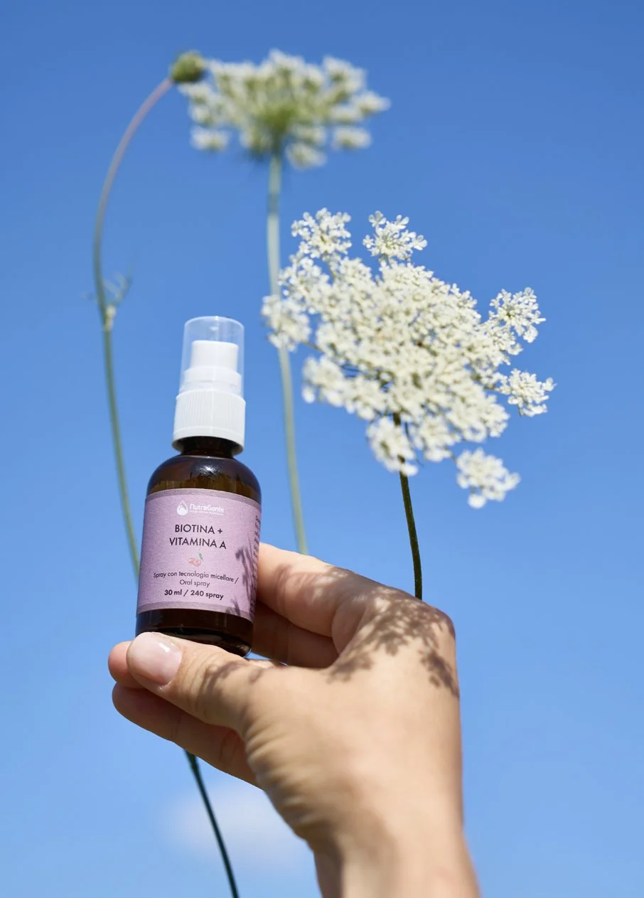 Hand holding a small bottle labeled 'Biotina +Vitamin A' against a background of white wildflowers and a clear blue sky.