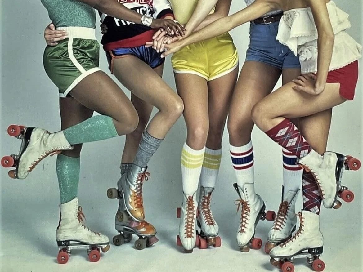 Groupe de femmes en immersion dans les années 80 debout sur des patins à roulettes, portant des vêtements colorés et des chaussettes longues, se tenant la main ou s'appuyant les unes sur les autres, dans une pose dynamique.