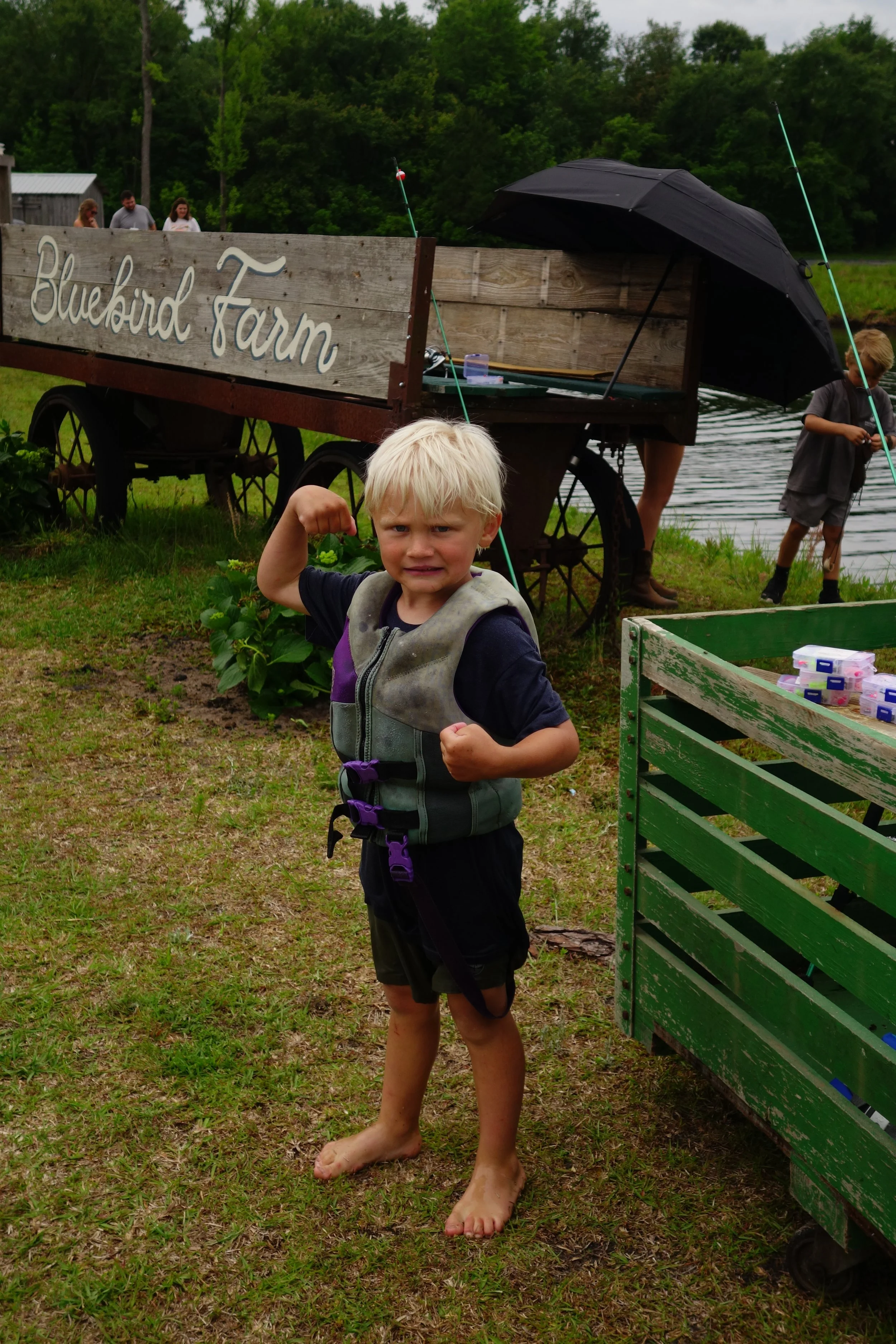 A young boy with blond hair, no shoes, and a life jacket, flexing his right arm in front of a farm sign that reads 'Bluebird Farm.' There is an umbrella, a fishing rod, and people near a body of water in the background.