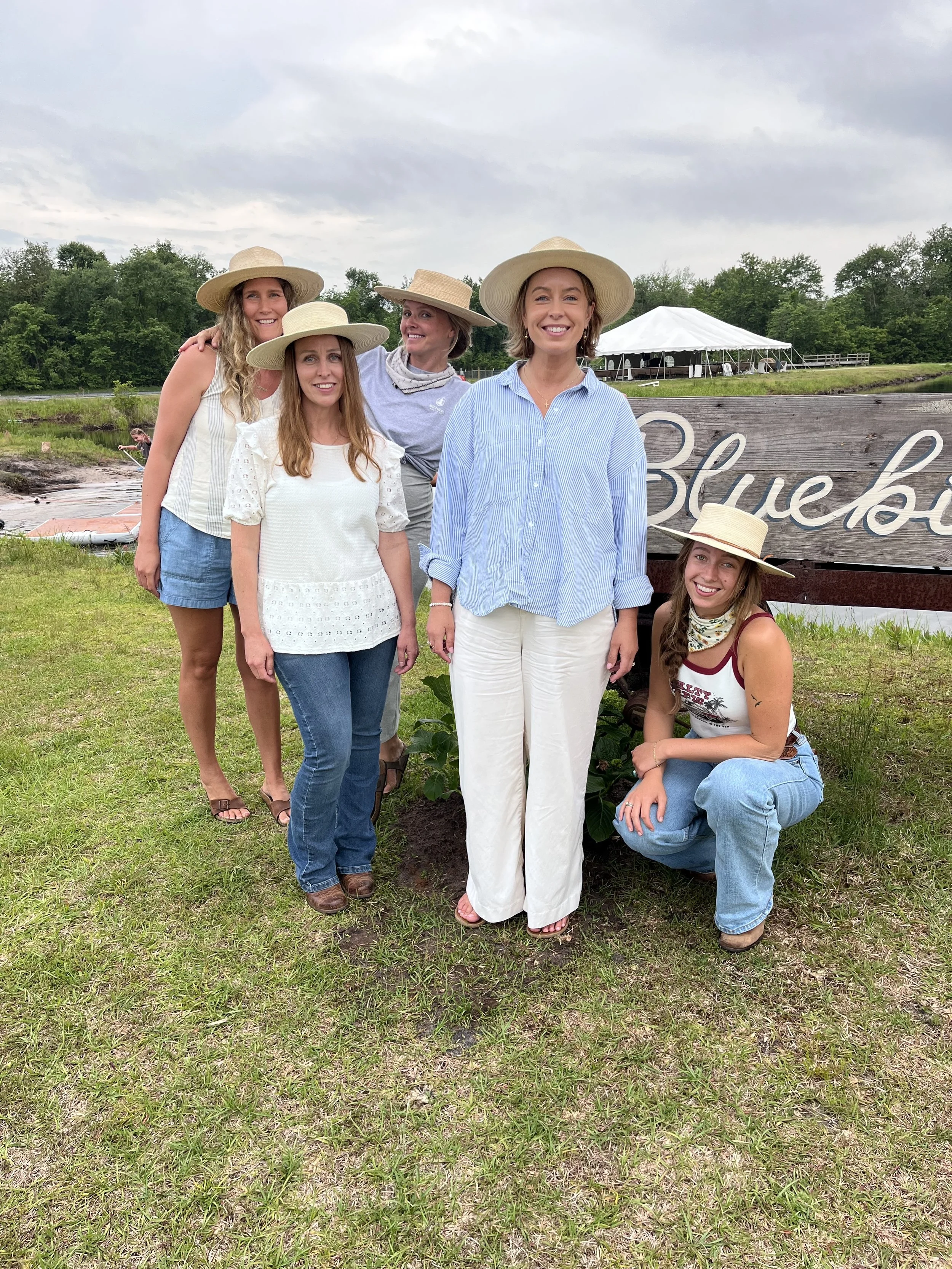 Group of six women outdoors on a cloudy day, wearing straw hats, standing and sitting near a large wooden sign with the partially visible word 'Blueb...'