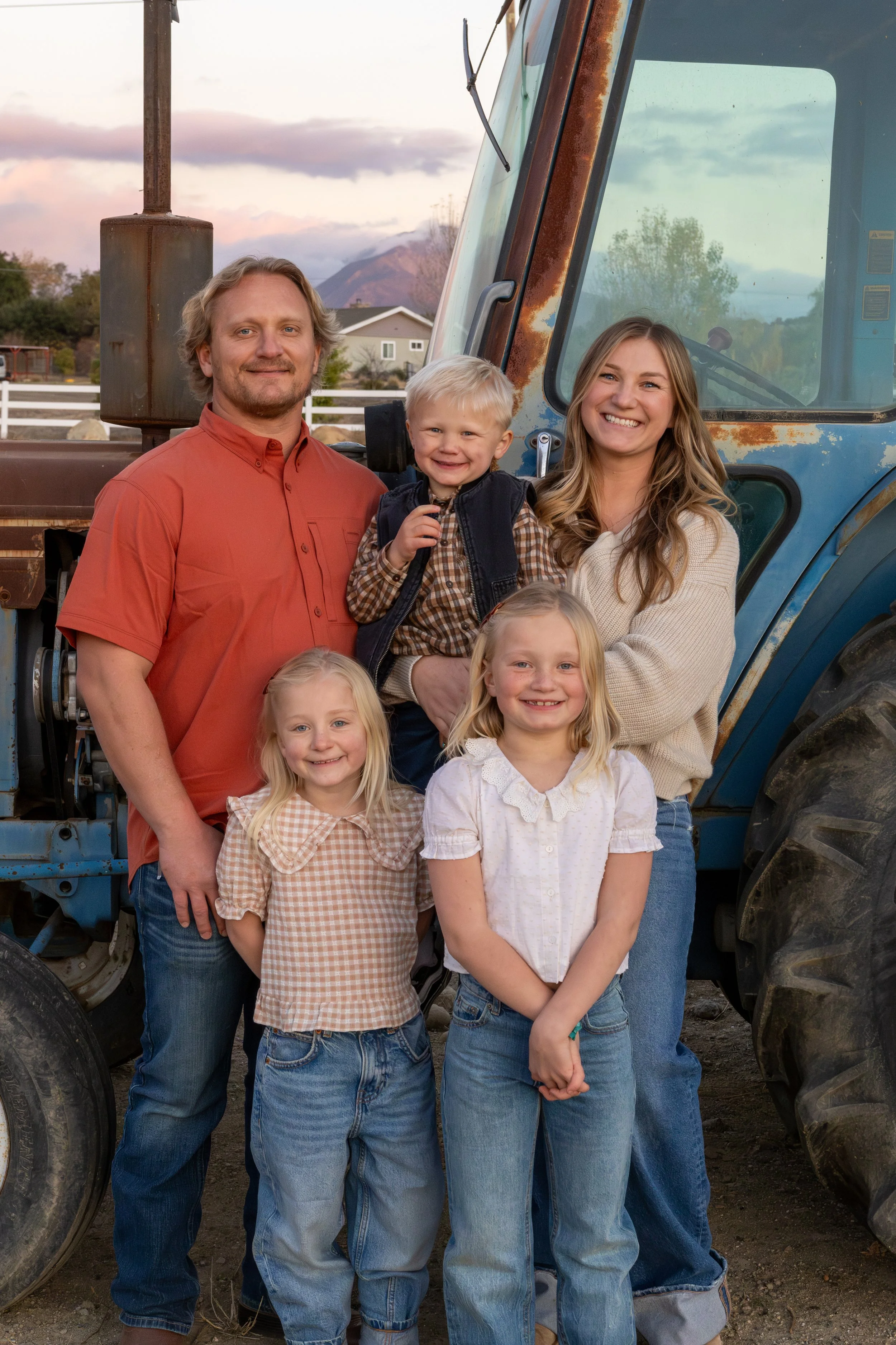 A family of six standing in front of a blue tractor outdoors during sunset, smiling at the camera.