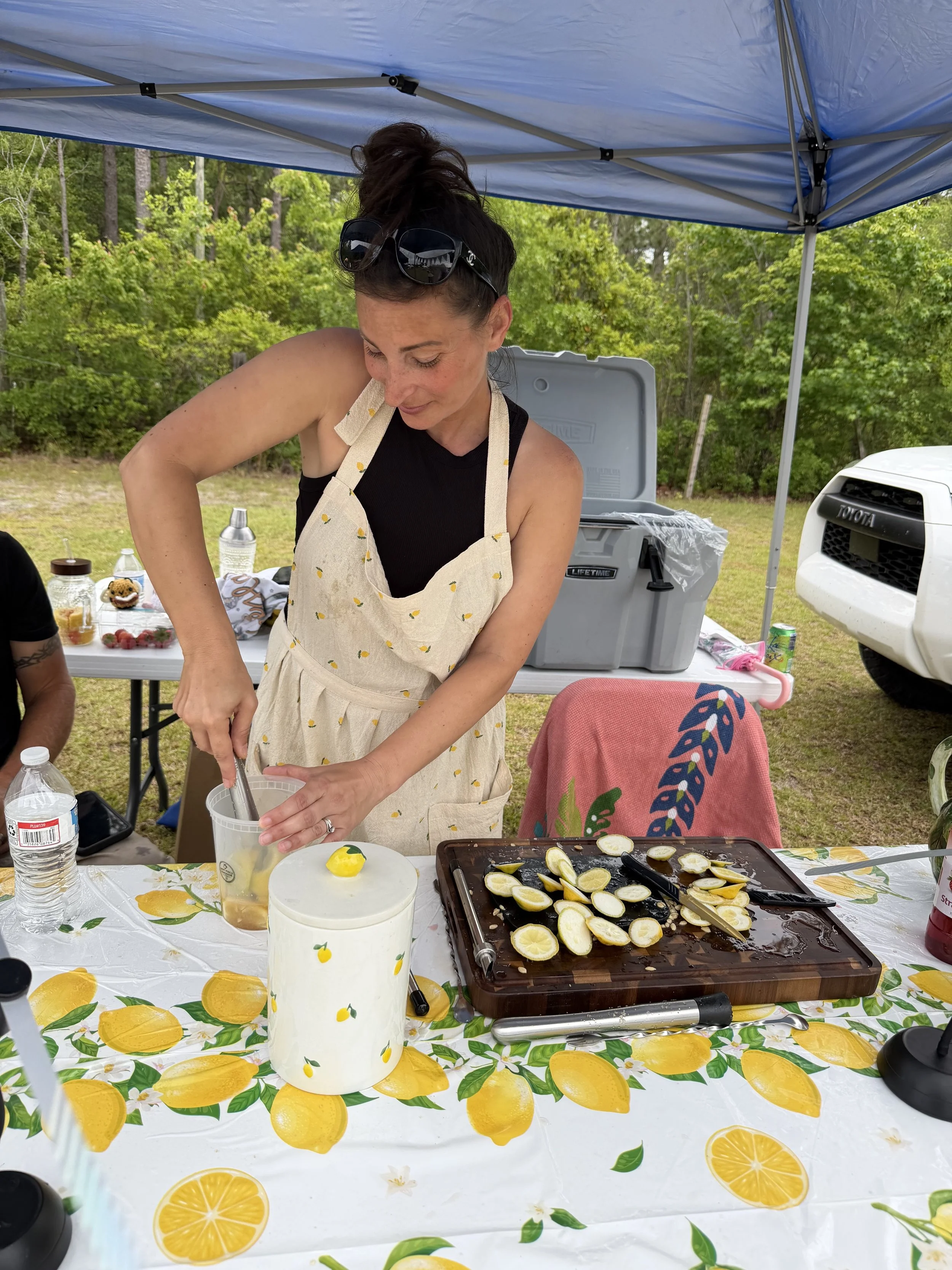 A woman wearing a lemon-print apron and black tank top is squeezing citrus fruit into a container at an outdoor gathering under a blue canopy. Various lemon slices and a lemon-themed tablecloth are visible on the table, with a cooler, a water bottle, and a car in the background.