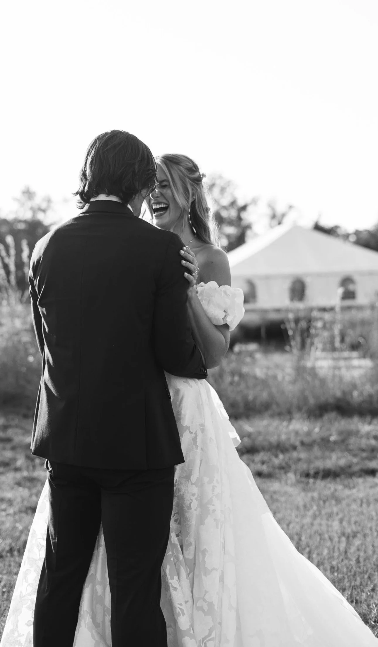 A black and white photo of a bride and groom dancing outdoors, smiling and laughing, with a tent and trees in the background.