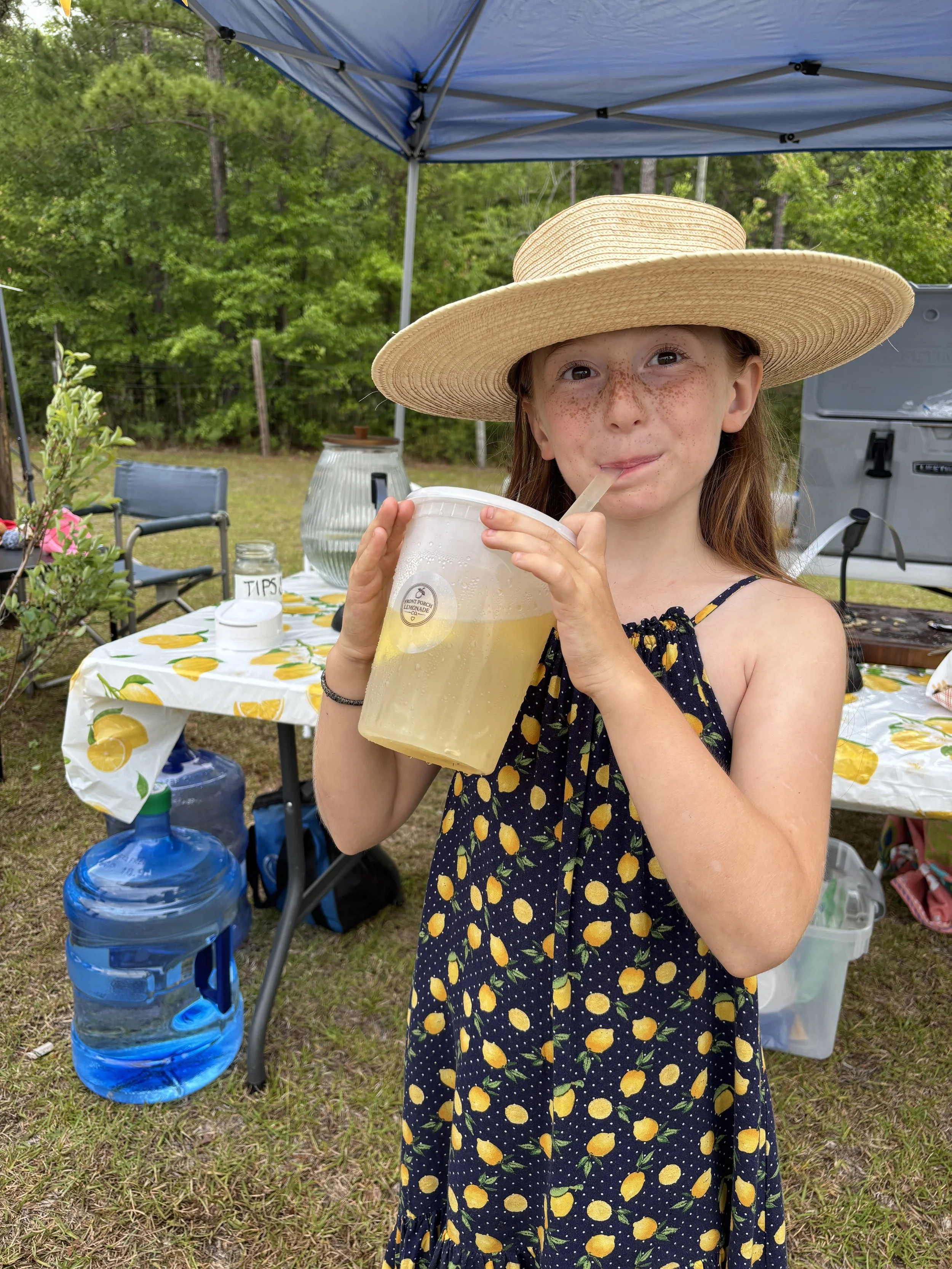 A young girl with freckles wearing a wide-brimmed straw hat and a navy dress with a lemon pattern, drinking lemonade from a large clear plastic cup with a straw at an outdoor gathering.