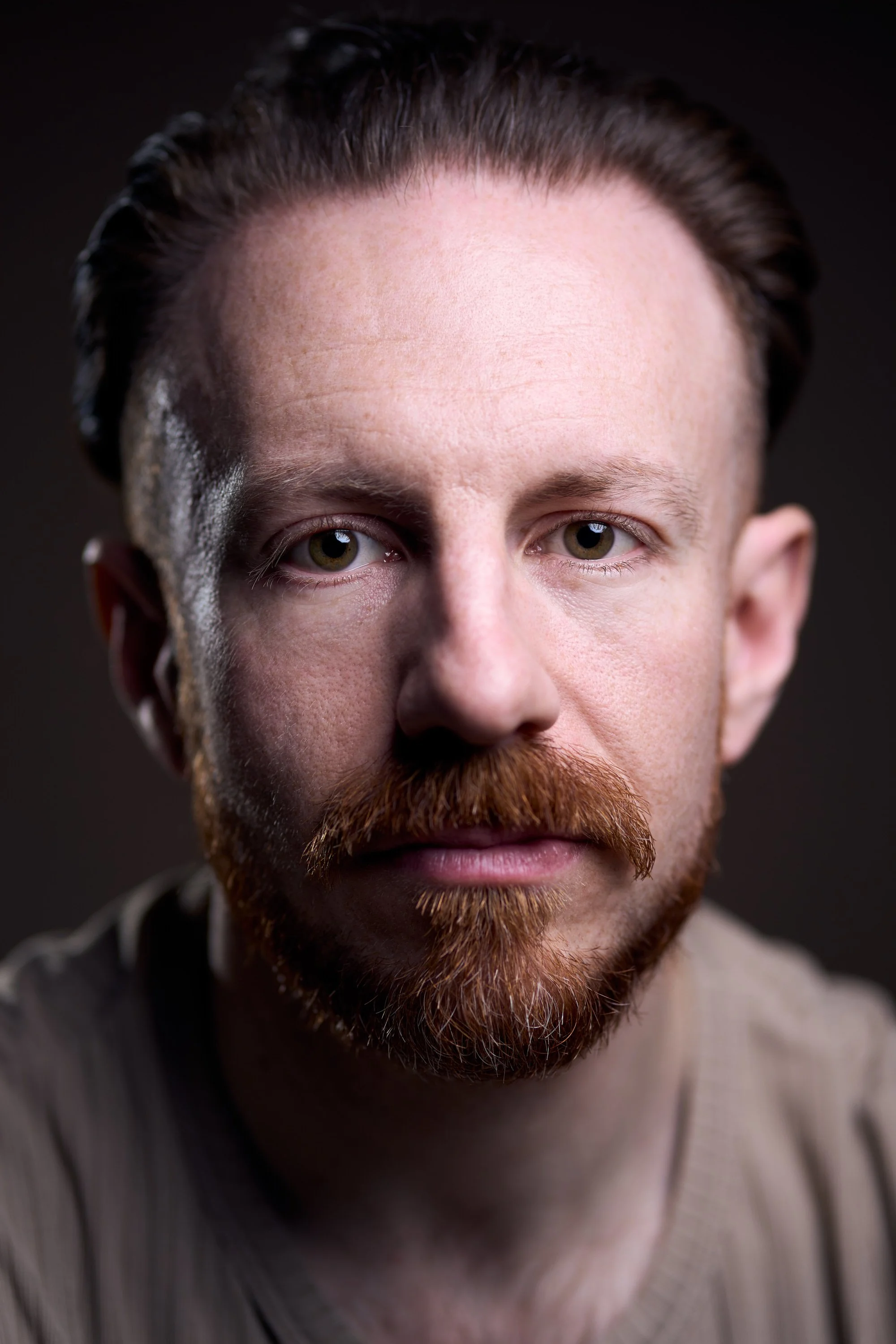 Close-up portrait of a man with light skin, reddish-brown hair, a matching beard and mustache, gazing directly at the camera against a dark background.